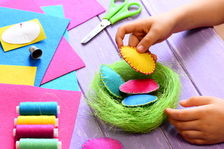 Small child does Easter decor. Child holds a felt egg in hand. Colorful felt Easter eggs and sisal nest, sewing tools and materials on a table. Simple and creative Easter decor ideaの写真素材