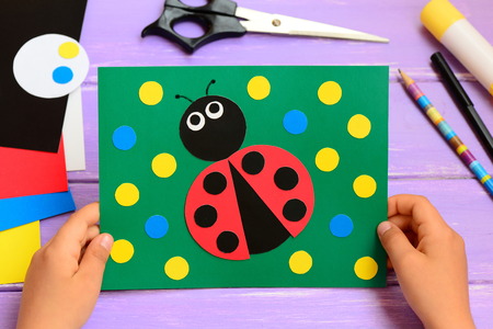Child holds a summer card in his hands. Child made a paper ladybug card. Stationery on a wooden table. Easy paper circle crafts for teaching kids to cut and glue. Developing kids motor skillsの写真素材
