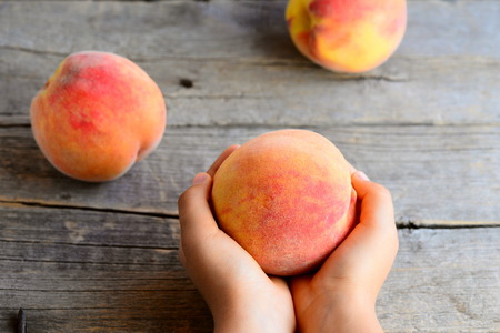 Small child holds a ripe peach in his hand. Juicy bright peaches on an old wooden table. Delicious and vitamin dessert for childrenの写真素材