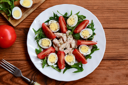 Healthy chicken vegetable salad. Homemade salad with fresh tomatoes, arugula, quail eggs, boiled chicken breast and spices on a white plate and a wooden table. Closeup. Top viewの写真素材