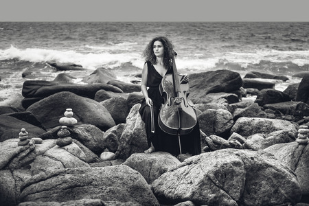beautiful young woman playing cello on stone beach at stormy weatherの写真素材