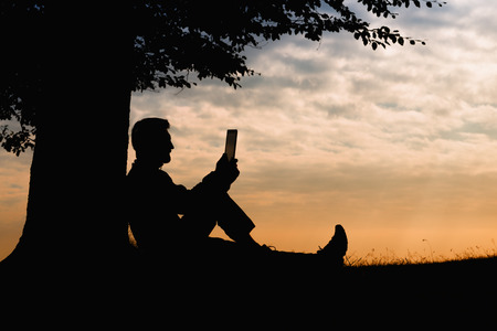 Man silhouette sitting under tree with book on cloudy day outdoorの写真素材