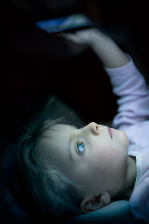 little child girl using tablet technology in bed by night at home. Serious or toughtful kid daughter in bedroom watching movie or reading or playing game. real people candid dark dreamy shotの写真素材