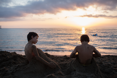 Young child boy and his friend or brother playing with sand at beach covering himself.Warm sunset light. Family summer travel vacations at sea or ocean.の写真素材