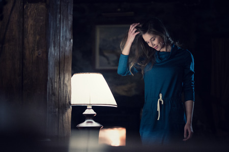 woman portrait wearing casual dress smiling and looking down near lamp it living room at rural home indoor. Europe Alps. Winter eveningの写真素材