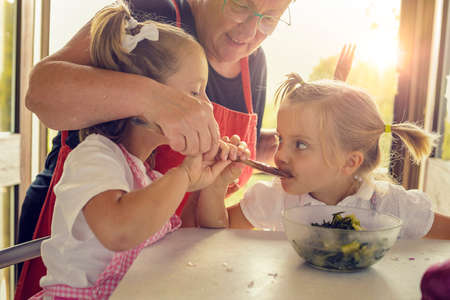 Two beautiful girls cooking cookies during morning light. family cooking, two kids learning by grandma mixing ingredients.Domestic life, family hobbiesの写真素材