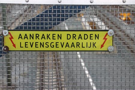 Warning sign with dutch text "aanrakeen draden levensgevaarlijk; which means touching wires is deadly, above electricity wires of trainのeditorial素材
