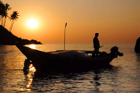 Silhouette of fisherman with nets in the sea on his boat at the dusk. Palm trees on the hill on backgroundの写真素材