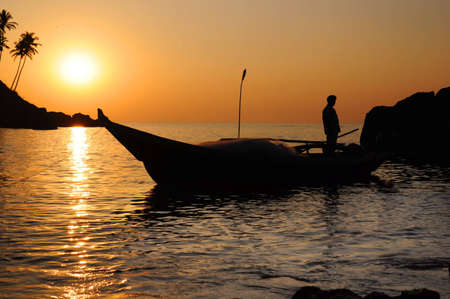 Silhouette of fisherman with nets in the sea on his boat at the dusk. Palm trees on the hill on backgroundの写真素材