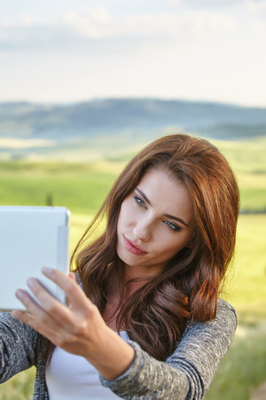 Beautiful brunette  woman  photographing selfie on a tablet on Tuscany hill backgroundの写真素材