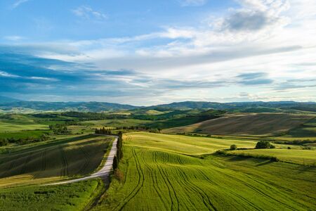 Picturesque Tuscany landscape with rolling hills, valleys, sunny fields, cypress trees along winding rural road, houses on a hill. Tourist visit in Tuscany の写真素材