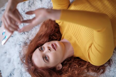 Smiling attractive young woman with beautiful long red hair lying in bed and taking selfieの写真素材