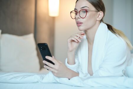 Beautiful young woman in bathrobe having rest on comfortable bed room after spa procedures. She is typing message on smartphone and smiling. Lady lies on comfortable bed and uses smartphone.の写真素材
