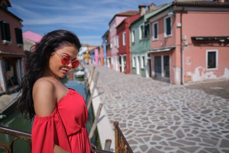 Young fashionable woman in romantic red  dress  with braid hairstyle, posing near colorful houses in Burano Island, Venice, Italyの写真素材