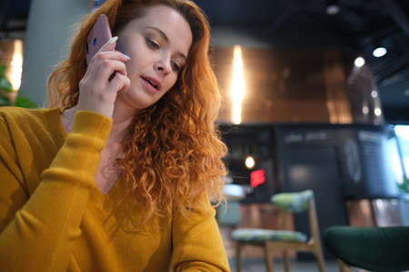 Young charming woman calling with cell telephone while sitting alone in coffee shop during free time, attractive female with cute smile having talking conversation with mobile phone while rest in cafeの写真素材
