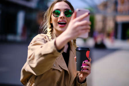 Closeup happy hipster girl doing selfie photo by mobile phone outdoors. Cheerful woman holding cellphone on city street. Laughing woman pulling faces with mobile phone in urban background.の写真素材