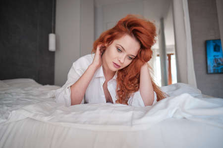 cute red hair woman in white shirt relax on the bed in her white roomの写真素材