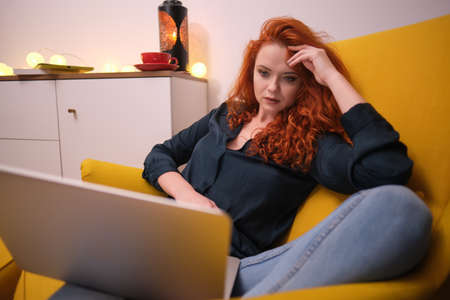 Nice beautiful lady with redhead curly hair work at the notebook sit down on the sofa at home - check on oline shops for cyber monday sales - technology woman concept for alternative office freelanceの写真素材