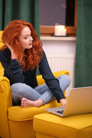 Nice beautiful lady with redhead curly hair work at the notebook sit down on the sofa at home - check on oline shops for cyber monday sales - technology woman concept for alternative office freelanceの写真素材