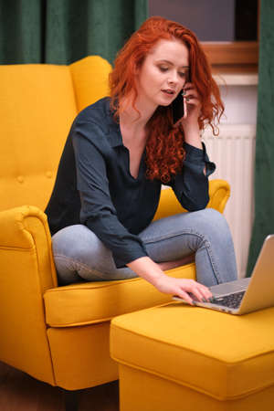 Nice beautiful lady with redhead curly hair work at the notebook sit down on the sofa at home - check on oline shops for cyber monday sales - technology woman concept for alternative office freelanceの写真素材