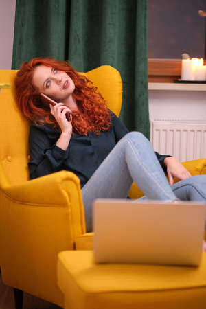 Nice beautiful lady with redhead curly hair work at the notebook sit down on the sofa at home - check on oline shops for cyber monday sales - technology woman concept for alternative office freelanceの写真素材