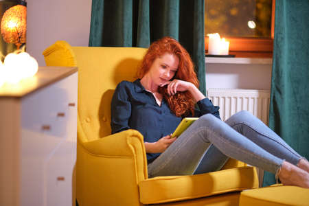 Woman using a tablet on line at home sitting on a yellow in the living room at home. Covid isolationの写真素材
