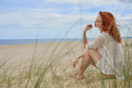 Smiling vivacious young woman relaxing on a beach sitting on the sand dunes overlooking the sea enjoying the sunshineの写真素材