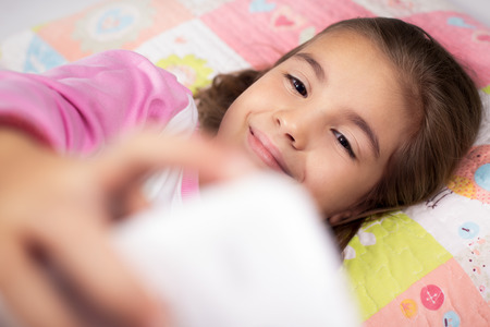 Little girl lying in bed with smartphone at homeの写真素材