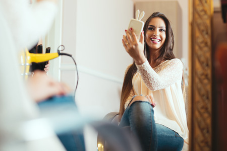 Happy young woman with smartphone taking mirror selfie at hair salonの写真素材