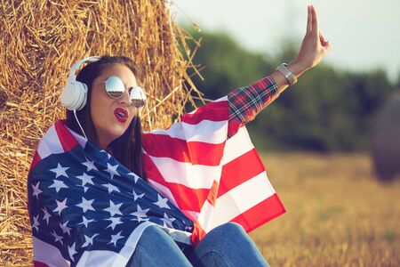 Beautiful girl sitting in the field, with a flag of the United States of America on her shoulders