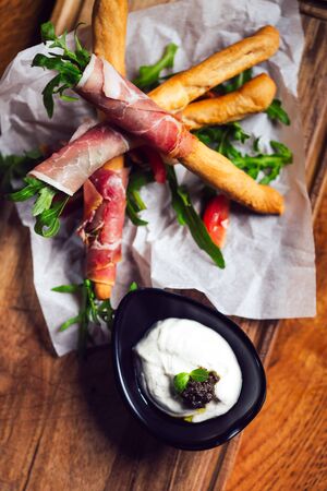 Bread sticks grissini with prosciutto ham on cutting board over wooden background.