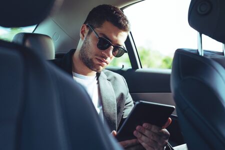 Young businessman sitting on back seat of car and touching digital tablet