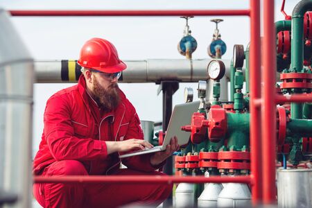 Engineer wearing safety helmet using laptop with oil refinery industry plant background.