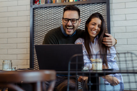 A couple sitting at cafe laughing cheerfully, looking at laptop screen, spending time with pleasure, drinking coffee.の写真素材