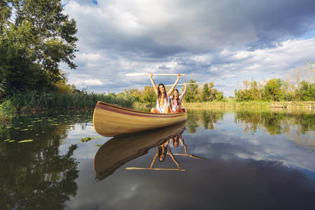 Two girls are paddling together in a wooden canoe on a calm, reflective lake surrounded by lush...