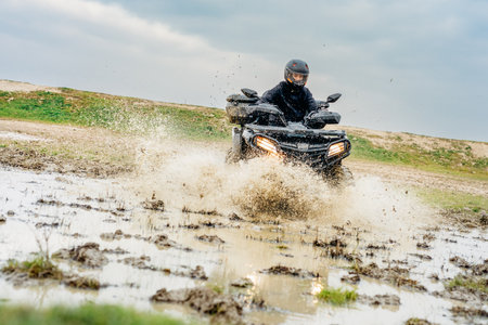 A rider wearing protective gear navigates an ATV through a muddy terrain, creating a dynamic splash, showcasing the thrill of off-road adventures.の写真素材