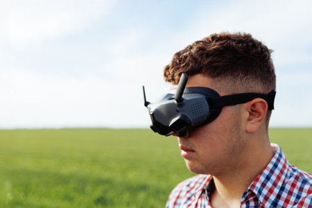 Short hair young man wears virtual reality headset with checkered shirt in open green field under blue skyの写真素材
