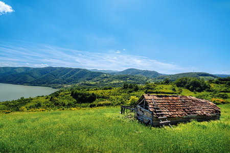 Scenic view of an old abandoned wooden hut in a green valley overlooking a river and rolling hills under a clear blue skyの写真素材
