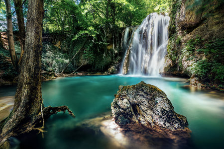 Stunning long exposure of waterfall cascading into vibrant turquoise pond, surrounded by lush green forestの写真素材