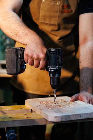 Carpenter wearing apron using cordless power drill making hole in wood plank in workshopの写真素材