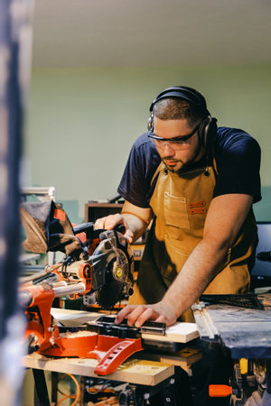 Carpenter wearing safety glasses and ear protection operating an electric saw while cutting a piece of wood in his workshopの写真素材