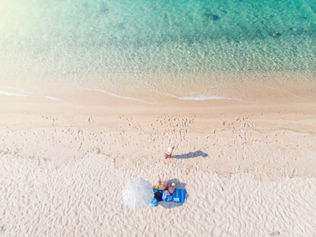 Drone aerial view from above of sandy beach, sea water and three-member family on towel next to sunshadeの写真素材