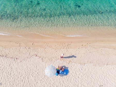 Drone aerial view from above of sandy beach, sea water and three-member family on towel next to sunshadeの写真素材