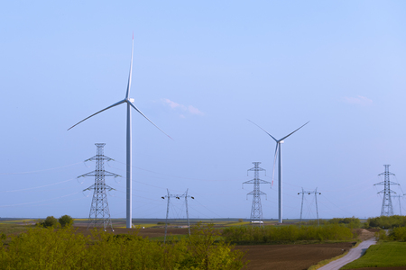 Two windmill turbines on agricultural field with large electricity towers in front, blue sky in background.の写真素材
