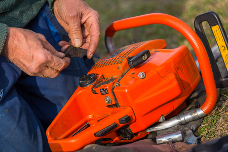 Old man in blue pants repair orange chainsaw placed on the ground with his bare hands.の写真素材