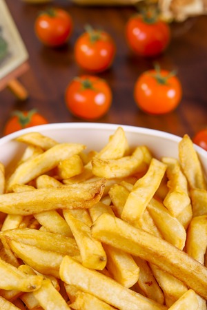 French fries in white bowl and tomato on wooden table.