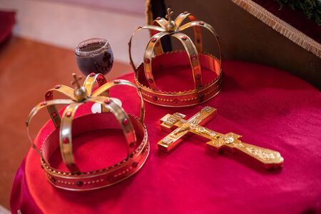 Cross, two crowns and glass of vine on red table prepared for wedding. Orthodox church wedding accessories.の写真素材