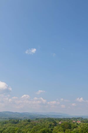 hilly landscape rich in tree vegetation with some houses and cloudy blue skyの写真素材