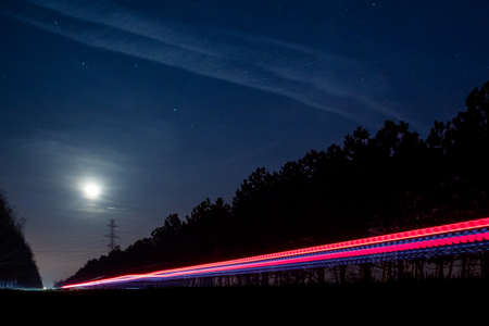 Light trails from vehicles traveling on rural road, surrounded by two rows of trees, late at night with moon on sky.の写真素材