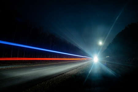 Light trails from vehicles traveling on rural road, surrounded by two rows of trees, late at night with moon on sky.の写真素材